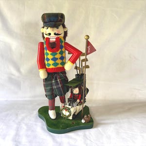 May include: A nutcracker figure dressed in a plaid cap, red sleeves, and a yellow and blue argyle sweater vest. The figure wears green plaid trousers and stands next to a miniature golf cart, a golf bag with clubs, and a red flag marked with 18.