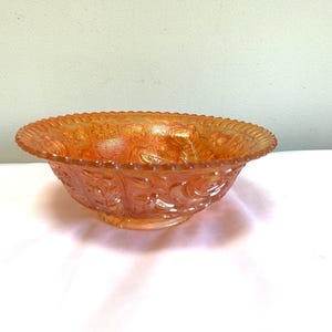 Puede incluir: Un cuenco de vidrio naranja iridiscente con un patrón floral decorativo. El cuenco tiene un borde festoneado y una base. El vidrio tiene una apariencia texturizada. Este cuenco vintage probablemente se usa para servir.