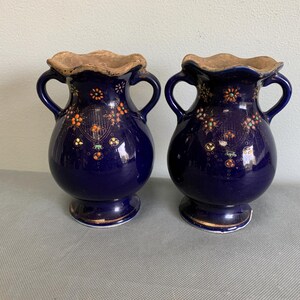 May include: A pair of deep blue ceramic vases with two handles each, featuring a ruffled, unglazed tan rim and intricate floral patterns in orange, green, and gold paint. The vases have a rounded body, a narrow neck, and a flared base.