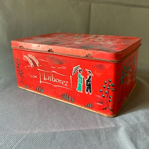 Può includere: Scatola di latta rossa con un design colorato che presenta due figure sotto un ombrello, un ponte e la parola "Labores".
