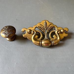 May include: A brass-toned metal drawer pull with a swinging handle and a matching round knob. Both pieces feature an embossed wheat stalk design. The pull has a decorative backplate with scrollwork details and a central mounting point for the handle.