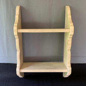 May include: A light-colored, two-tiered wooden shelf with decorative side supports. The shelf is made of unfinished wood and is designed to be mounted on a wall. The shelf is approximately 24 inches wide and 20 inches tall.