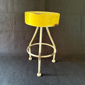 May include: A small stool featuring a round, bright yellow seat with visible signs of wear and rust along the edges. The base consists of three thin, off-white metal legs connected by a circular footrest ring, also showing patches of rust.