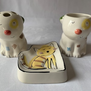 以下が含まれることがあります： 3つのセラミックバスルームアクセサリーのセット。緑色の耳、黄色の斑点、赤い鼻を持つ犬の形をした容器が2つ。黄色の猫のデザインの猫の形をした石鹸皿。セットは白で、カラフルなアクセントが付いています。