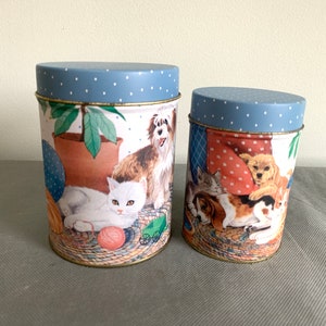 Op de afbeelding: Twee blauwe en witte metalen blikken met stippen met afbeeldingen van katten en honden. De grotere blik heeft een witte kat met een bol wol en een bruin-witte hond. De kleinere blik heeft een bruin-witte hond, een zwart-witte kat en een bruin-witte kat.