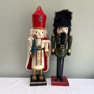 May include: Two wooden nutcrackers standing side by side. One is dressed as a bishop in a red mitre with a blue cross, white robe, and red cape, holding a small green tree. The other is a soldier in a black bearskin hat and green uniform.