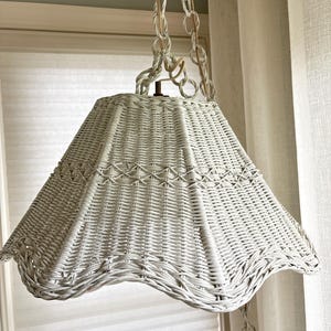 May include: A white wicker pendant light fixture with a flared, scalloped bottom edge and a woven lattice pattern. The lamp shade is suspended by a matching white wicker chain that connects to a central metal hook at the top of the fixture.