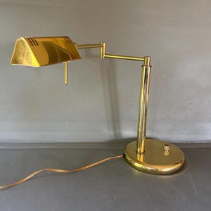 May include: A polished brass desk lamp featuring a wide, rectangular shade with ventilation slits, an adjustable articulated arm, and a circular base. The lamp includes a small control knob on the base and a thin, light-colored electrical power cord.