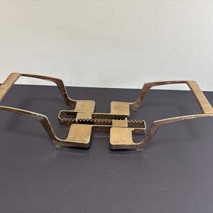 May include: An expandable brass trivet featuring a central interlocking mechanism with a serrated adjustment bar and two wide, angled handles on either side. The metallic surface has a brushed finish with visible horizontal lines on the handle grips.