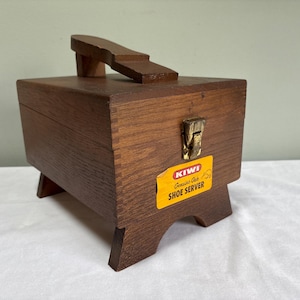 Peut inclure: Une boîte à cirer les chaussures en bois vintage, de couleur marron, avec une poignée et un couvercle à charnière. La boîte est rectangulaire et repose sur quatre petits pieds. Une étiquette sur le devant indique "KIWI Shoe Server".