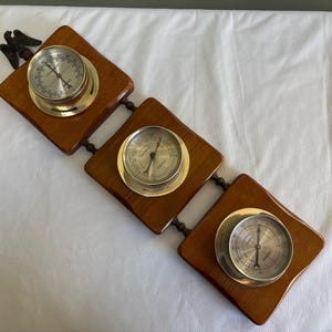 May include: A vintage weather station featuring three round gauges with silver faces and gold-colored rims, set on diamond-shaped wooden plaques. The plaques are connected by a chain and have a dark brown finish. An ornate metal bird decorates the top plaque.