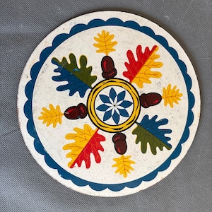 May include: Round, white coaster with a blue scalloped edge. The coaster features a central floral design surrounded by colorful leaves and acorns. The leaves are in shades of yellow, red, and green. The text "ZOOK" is visible on the edge.
