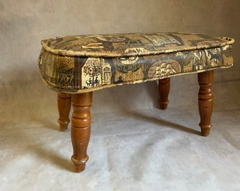 May include: An oval-shaped footstool with a patterned top featuring a brown and tan landscape design. The stool has four turned wooden legs. The fabric is bordered with a light brown trim. A vintage-style accent piece.