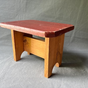 May include: A small, rectangular wooden stool with a red-painted top and natural wood legs. The stool is approximately 12 inches long, 8 inches wide, and 10 inches tall. The design is simple and rustic, with visible wood grain.