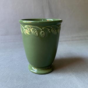 May include: A green ceramic vase with a pedestal base and a flared top. The rim is decorated with a raised leaf pattern. The vase has a smooth, matte finish and is approximately 6 inches tall.