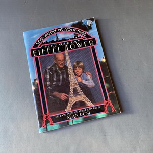 以下が含まれることがあります： エッフェル塔の模型を組み立てている2人の写真が表紙の書籍。書籍のタイトルは「The World at Your Feet: Build Your Own Eiffel Tower」、著者はアラン・ローズです。