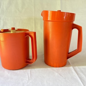 Peut inclure: Deux récipients en plastique orange vif avec poignées. Le récipient le plus court à gauche présente un couvercle plat avec un petit bouton central. La carafe plus haute à droite possède un couvercle orange assorti et une poignée latérale.
