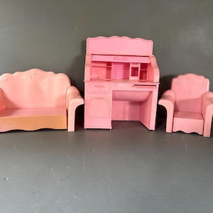 May include: A set of three pink plastic dollhouse furniture pieces including a sofa with a curved back, a roll-top desk with integrated shelving and drawers, and a matching armchair with a scalloped backrest, all featuring a smooth finish.