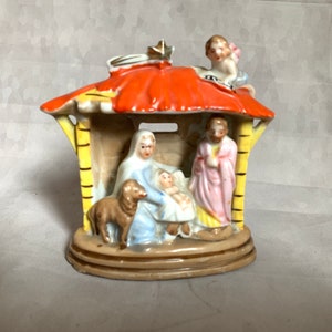 May include: A ceramic nativity scene figurine featuring a bright orange roof with a gold star, a small angel, and a stable structure. Inside, figures of a person in blue, a person in pink, a baby, and a brown sheep are depicted on a tan base.