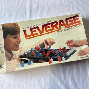May include: Vintage Milton Bradley "Leverage" board game box. The box features the game's title in large orange letters, with the tagline "A Game of Strategy and Suspense." The game board is black with red, blue, and white game pieces.