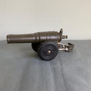 Könnte beinhalten: Eine Spielzeugkanone aus Metall mit dunkelgrüner Lackierung. Die Kanone hat zwei schwarze Gummireifen und eine Metallbasis. Die Kanone steht auf einer grauen Oberfläche.