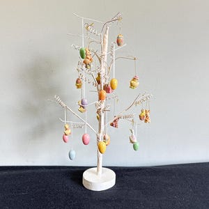 May include: A white Easter tree with decorative hanging ornaments. The ornaments include colorful eggs and small figurines of bunnies and chicks. The tree is on a white circular base and has a light-colored background.