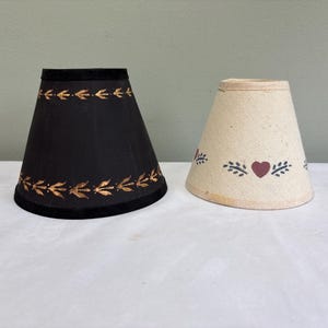 May include: Two conical lamp shades. The left shade is black with gold-colored leaf patterns along the top and bottom edges. The right shade is off-white with a repeating pattern of small dark red hearts and green leaf sprigs around the center.