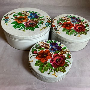 Peut inclure: Ensemble de trois boîtes de rangement vintage en métal blanc. Chaque boîte présente un motif floral au point de croix sur le couvercle avec des fleurs rouges, orange, violettes et bleues. Les boîtes sont ovales et rondes, avec des côtés côtelés.