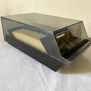 May include: A black Rolodex rotary card file organizer featuring a translucent dark gray plastic cover. The unit contains a series of cream-colored index cards arranged in a row, with the brand name Rolodex printed in white on a black label.