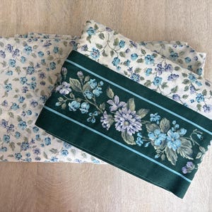 May include: A folded fabric sheet featuring a white background with a small-scale blue and purple floral pattern. A wide, dark green horizontal band runs across the fabric, decorated with a larger floral print in shades of blue and purple.