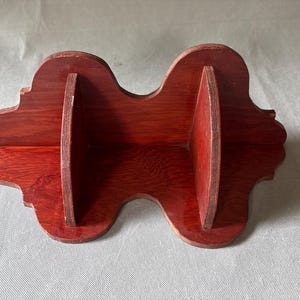 Op de afbeelding: Een houten plank met een rijke, roodbruine afwerking. De plank heeft een uniek, gebogen ontwerp met twee verticale verdelers, waardoor drie afzonderlijke displayruimtes ontstaan. De randen zijn subtiel geschulpt, wat een decoratief tintje geeft aan dit woondecoratie-item.