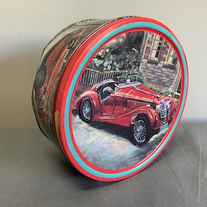 May include: A round, red tin with a vintage car design. The tin features a red car on a road, with a house and trees in the background. The rim has a teal and red stripe.