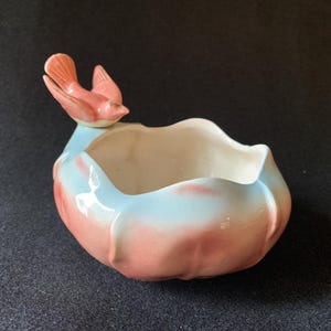 May include: A ceramic planter in a tulip shape, painted in shades of pink and light blue. A pink bird with gold accents perches on the rim. The interior is white. A decorative piece for the home.
