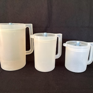 May include: Three translucent plastic pitchers of varying sizes with lids and handles. The pitchers are light in color and appear to be empty. The background is a solid black.
