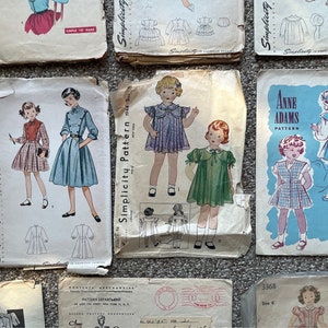 May include: A collection of vintage Simplicity sewing patterns for children's clothing. The patterns feature illustrations of dresses and outfits, with text including "Simplicity Printed Pattern 25c" and "Anne Adams Pattern".