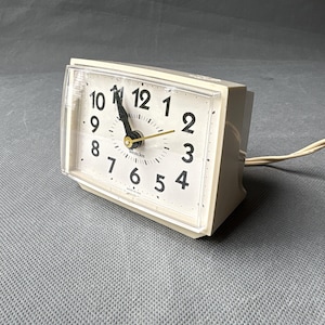 Pode incluir: Um despertador elétrico vintage creme com uma capa de plástico transparente. O mostrador do relógio tem algarismos e ponteiros pretos, com um ponteiro de segundos dourado. O relógio diz "Westclox" e tem um cabo de alimentação.