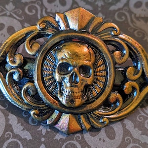 Puede incluir: Una pinza para el pelo de metal dorado con un diseño ornamentado que presenta una calavera en el centro. La calavera está detallada con una superficie texturizada y tiene un esquema de color negro y dorado.