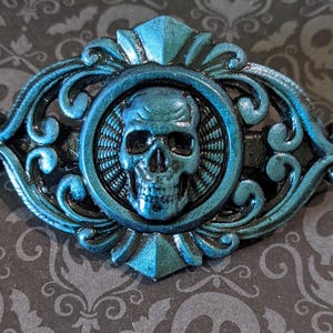 Op de afbeelding: Een tealblauwe metalen haarspeld met een decoratief schedelontwerp. De schedel bevindt zich in het midden van een cirkelvormig frame met een gedetailleerd ontwerp. De speld is versierd en heeft een vintage stijl.
