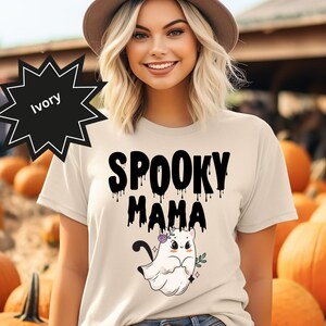 Könnte beinhalten: Elfenbeinfarbenes T-Shirt mit dem Text "SPOOKY MAMA" in schwarzer Tropfschrift. Unter dem Text befindet sich ein Cartoon-Geist mit Blumenkrone. Der Hintergrund zeigt Kürbisse und eine Person mit Hut.