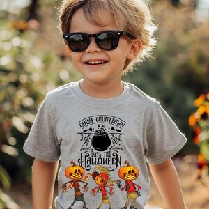 Könnte beinhalten: Ein graues T-Shirt mit einem Halloween-Motiv. Der Aufdruck zeigt den Text "Candy Countdown Days Till Halloween" über drei Cartoon-Vogelscheuchen mit Kürbisköpfen. Das Kind trägt eine schwarze Sonnenbrille und ein schwarzes Armband.