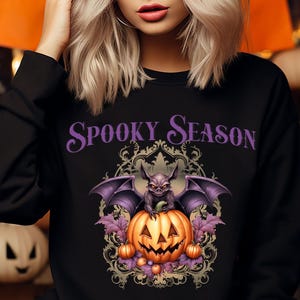 Könnte beinhalten: Schwarzes Sweatshirt mit dem Text "Spooky Season" in Lila. Die Grafik zeigt eine Fledermaus, einen Kürbis und andere Halloween-Elemente. Der Hintergrund umfasst orangefarbene Dekorationen und Kürbisse.