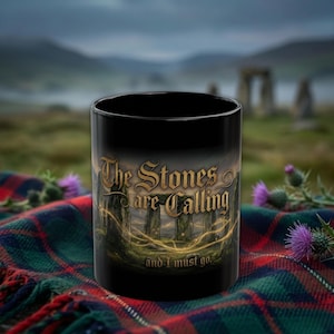 Könnte beinhalten: Schwarze Keramik-Tasse mit dem Text "The Stones are Calling and I must go." Die Tasse zeigt ein Landschaftsdesign mit alten Steinen und einem goldenen Schein. Die Tasse steht auf einer karierten Decke.