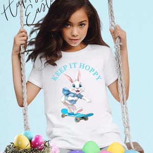 Könnte beinhalten: Ein weißes T-Shirt mit einem blau-weißen Hasen, der Skateboard fährt, und dem Text "Keep It Hoppy". Das Mädchen trägt das Shirt und sitzt auf einer Schaukel. Im Vordergrund befinden sich Ostereier in einem Nest.