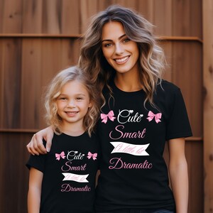 Könnte beinhalten: Zwei schwarze T-Shirts mit dem Text "Cute Smart A little bit Dramatic" in Weiß und Rosa. Die Shirts haben rosa Schleifenakzente. Die Shirts werden von einer Frau und einem jungen Mädchen getragen.