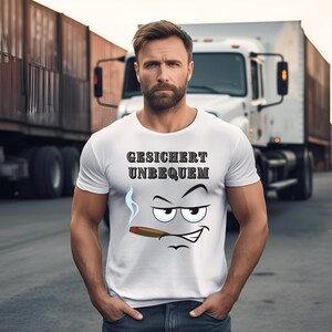 Op de afbeelding: Wit t-shirt met de tekst "GESICHERT UNBEQUEM" en een cartoongezicht dat een sigaar rookt. Het gezicht heeft een humeurige uitdrukking met een grijns. Op de achtergrond zijn een vrachtwagen en aanhangers te zien.