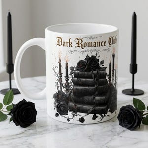 Puede incluir: Taza de cerámica blanca con el texto "Dark Romance Club" en letras doradas. La taza presenta un diseño gótico con rosas negras, libros apilados y velas encendidas. Dos velas negras y rosas están sobre la mesa.