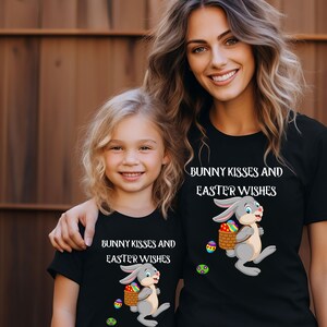 Könnte beinhalten: Zwei Personen tragen schwarze T-Shirts mit einem Hasen, der einen Korb mit Ostereiern hält, und dem Text "Bunny Kisses and Easter Wishes".