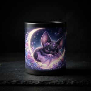 Könnte beinhalten: Ein schwarzer Keramikbecher mit einer grafischen Illustration einer kleinen Fledermaus mit geschlossenen Augen, die eine juwelenbesetzte Krone trägt. Die Fledermaus ruht in einem leuchtenden Halbmond, umgeben von Rosen und Kristallen.