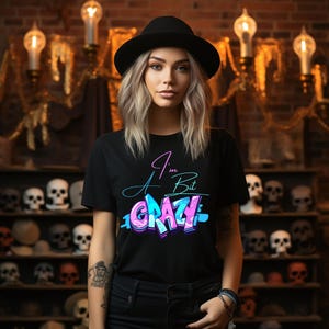 Könnte beinhalten: Schwarzes T-Shirt mit dem Text "I'm A Bit CRAZY" in einem farbenfrohen Graffiti-Stil. Das Model trägt einen schwarzen Hut. Der Hintergrund zeigt eine Backsteinmauer und Regale mit Totenköpfen.