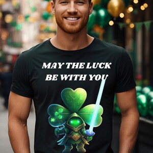 Könnte beinhalten: Ein schwarzes T-Shirt mit einem grünen vierblättrigen Kleeblatt und einem Lichtschwert. Der Text "Möge das Glück mit dir sein" ist auf dem Shirt gedruckt.